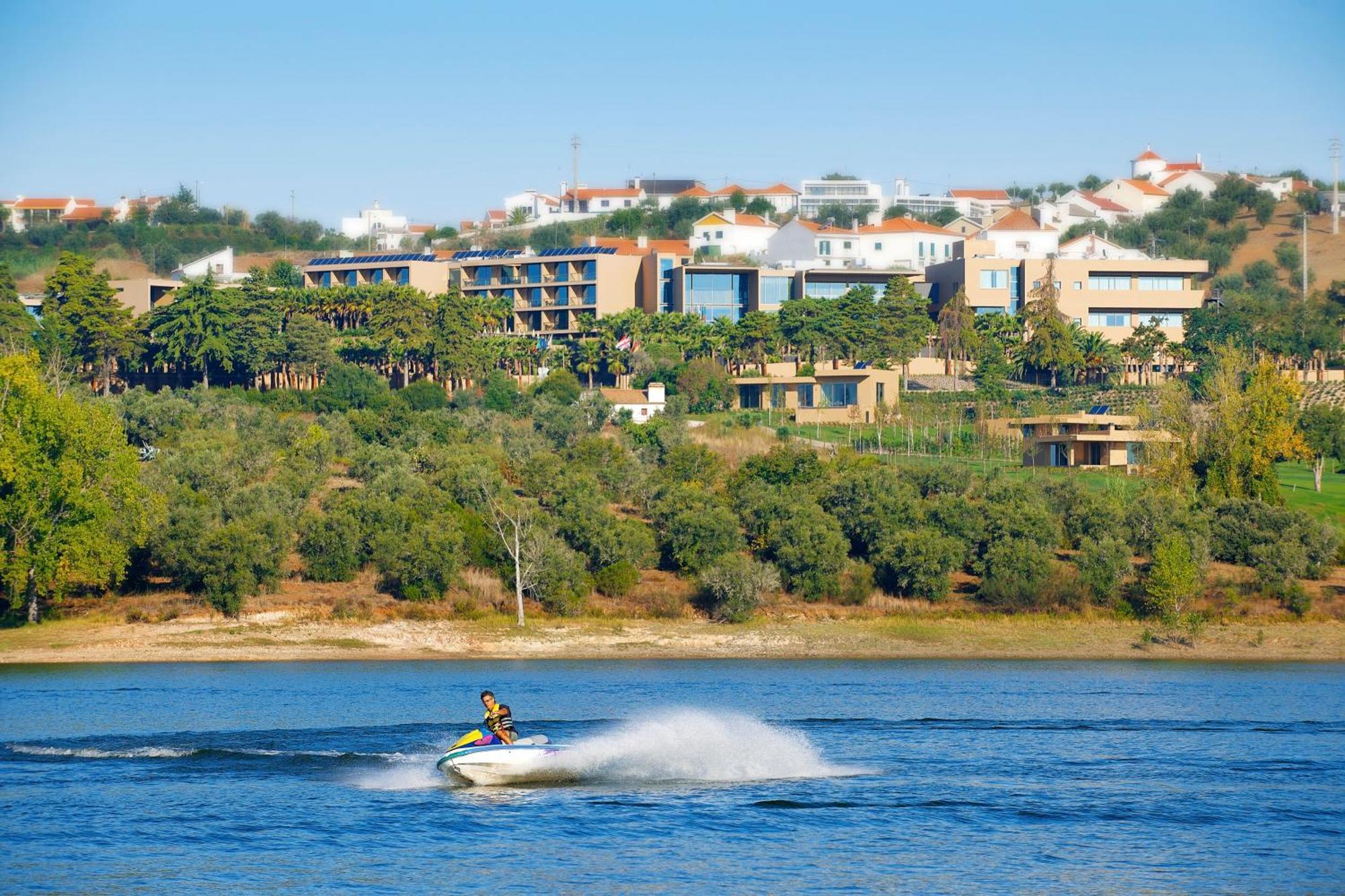 拉戈芒塔格别墅瑙酒店 蒙塔吉尔 外观 照片 The photo shows a person riding a jet ski on a body of water, creating a spray as they speed across the surface. In the background, there are modern buildings and a hillside with trees and greenery. The sky is clear and blue, suggesting a bright and 